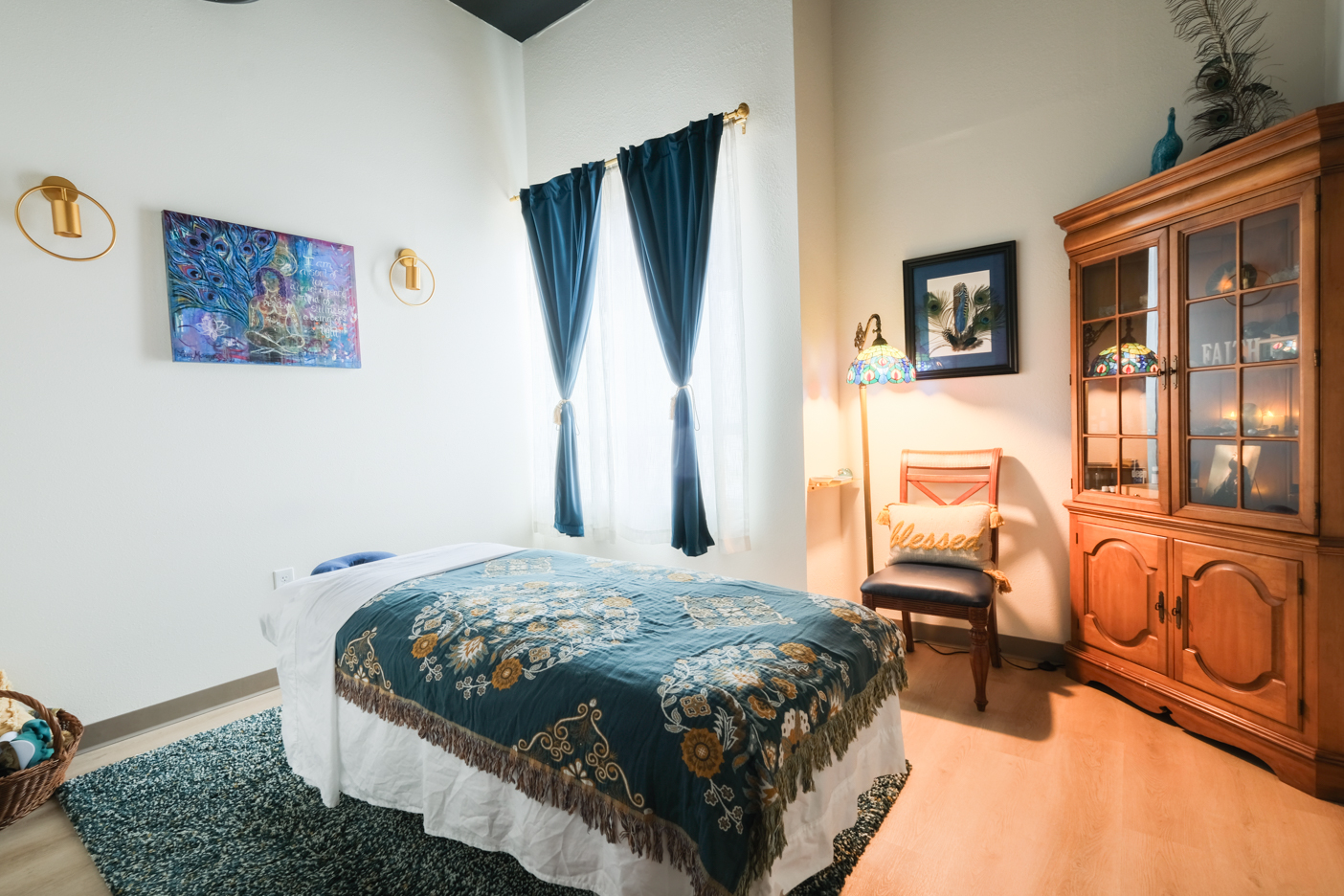 Warmly lit massage room at Flora Integrated Wellness in Escondido with a treatment table, blue curtains, peacock artwork, and a wooden hutch